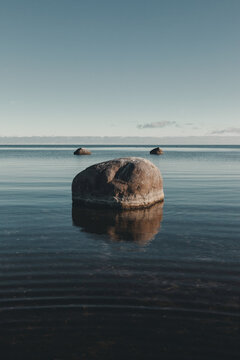 View of a large boulder resting in the calm, clear waters off the coast of Vormsi, with smaller rocks visible on the horizon under a vast blue sky in Laane County, Estonia.