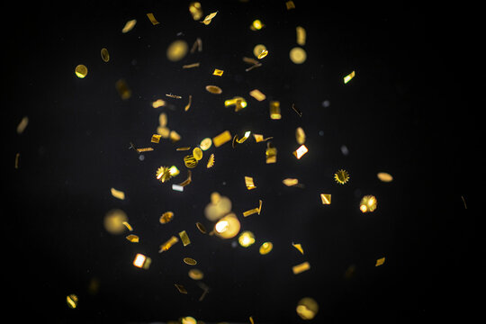 Shimmering golden glitter confetti pieces in various shapes including circles squares and starbursts falling through the dark air creating a festive and celebratory atmosphere of joy