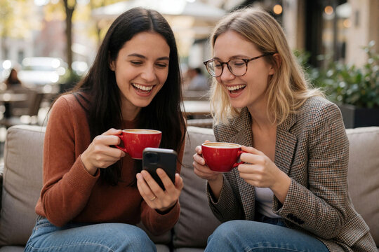 Two women friends laugh looking at a smartphone holding coffee cups. They sit on a couch outdoors, sharing a happy moment. Casual conversation and connection over coffee and tech.