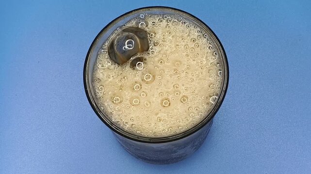 Pouring or filling sweet fizzy soft soda liquid beverage inside glass filled with ice cubes. Unhealthy non-alcoholic mocktail summer cold drink foam and bubbles closeup top view.