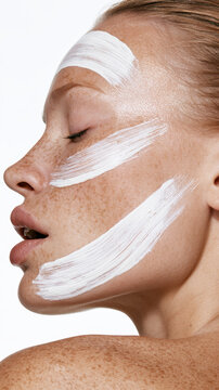 Woman's closed eyes focusing on skincare, applying a thick white cosmetic product on her forehead, cheek, and chin, demonstrating a beauty routine for healthy skin