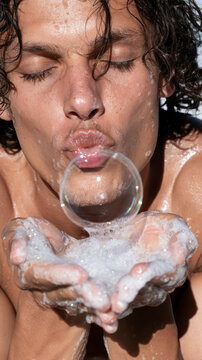 Wet man with closed eyes blowing soap bubble from foamy hands, conveying concepts of freshness, cleanliness, hygiene, and enjoyment of personal care for summer skin rejuvenation