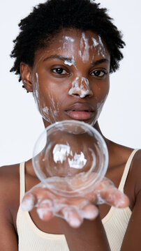 Young black woman washing face with cleanser, foam covering skin, holding a delicate glass bubble in her outstretched hand, representing purity, freshness, and effective skincare