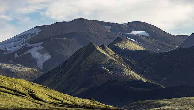 View of mossy green volcanic mountains with patches of snow under a dramatic sky in Iceland.