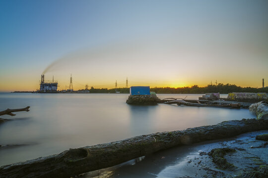 View of a sunset over the coast with an industrial power plant in the distance, calm water, and driftwood on the shore in San Pedro de Macoris, San Pedro de Macoris, Dominican Republic.