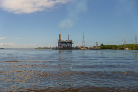 View of an industrial power plant on the water with smoke, power lines, and calm sea under a clear blue sky in San Pedro de Macoris, San Pedro de Macoris, Dominican Republic.