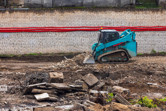 View of a teal skid-steer loader working on a construction site with piles of dirt and rubble in front of a brick wall in Vilnius, Vilnius County, Lithuania.