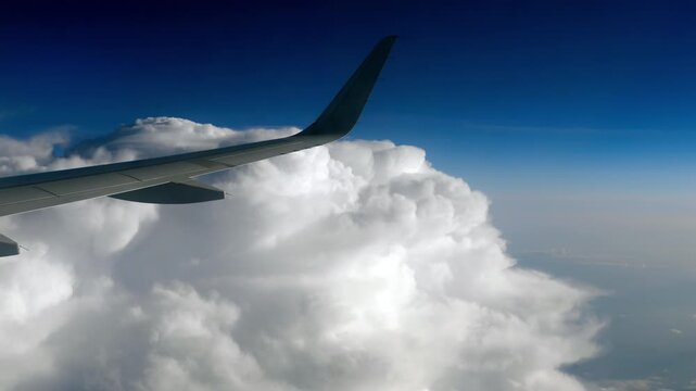 Airplane wing flying above massive white cumulus clouds under a deep blue sky in bright daylight, shot in real time from a passenger window at high altitude.