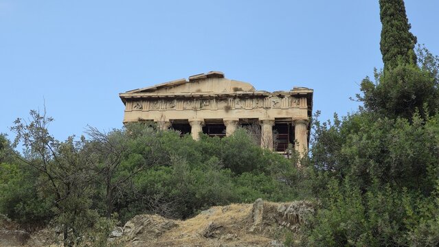 Athens, Greece - 8 July 2026. Temple of Hephaestus in the Ancient Agora seen above olive trees and shrubs, with Doric architectural columns, interior scaffolding, a cypress tree, and clear sky.