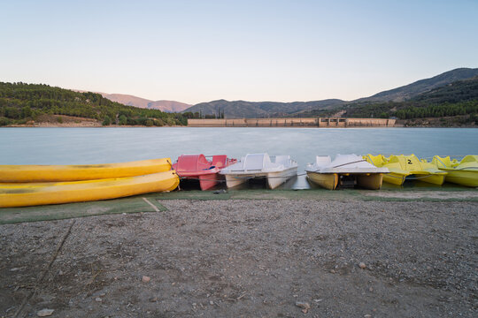 Pedal boats and kayaks lined up on shore of beznar reservoir with a dam and mountains in background