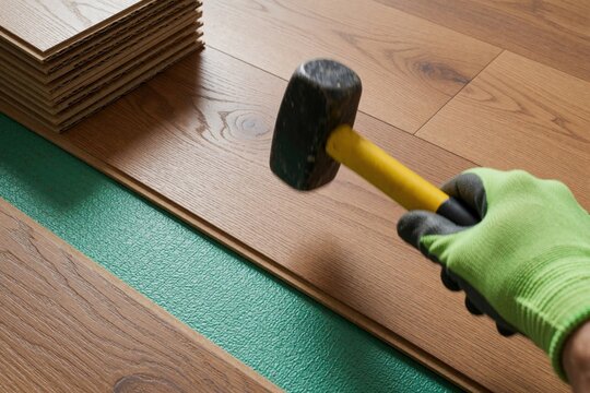 Worker installs brown wooden floor panels inside residential house using rubber mallet tool. Handyman wears protective glove tapping planks together on green underlay base. Person performs interior