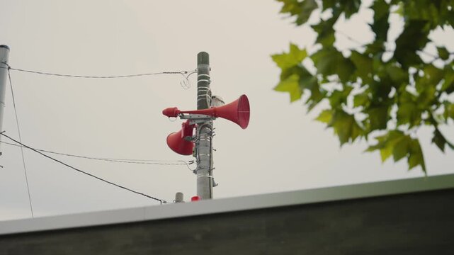 Red loudspeakers on a utility pole used for public announcements and emergency alerts in a Japanese city, with green tree leaves and a cloudy sky visible in the background in city or town