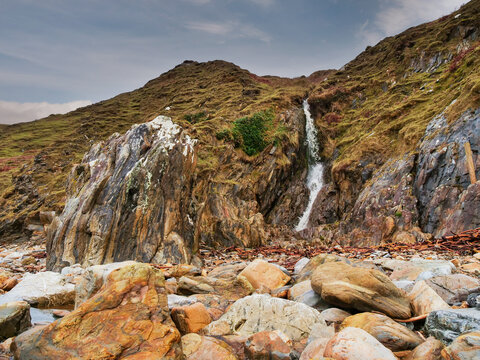 Small waterfall flows down mossy cliffside. Pebble beach stretches beneath rocky outcrop. Wildlife-free nature scene with greenery and water. No animals or plants identified in image. Nature beauty.