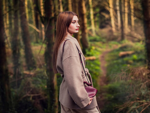 Young woman walks through autumn forest, carrying burgundy bag. Trees show vibrant fall colors, leaves cover ground. Image captures serene forest park walk. Stylish young lady outdoors. Elegant girl