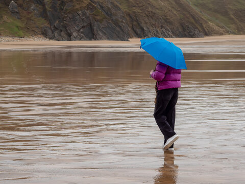 Raindrops patter on a vivid blue umbrella. Figure in purple puffer jacket stands alone. Wet sand reflects the gray, stormy sky. Cliffs loom in the background, wild and imposing. Travel and tourism.
