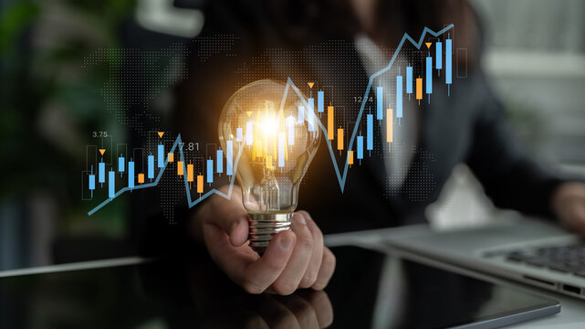 A business professional holds a glowing light bulb symbolizing innovation against financial charts, illustrating the relationship between creativity and market growth. Gantry