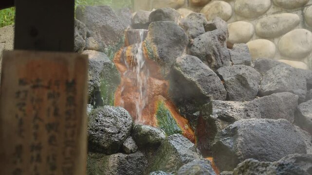 Steaming mineral rich water from a natural onsen cascading down colorful volcanic rocks, a traditional hot spring source for bathing and wellness located in nature of Hokkaido in a public foot bath
