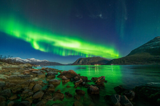 View of the northern lights illuminating the night sky over a rocky arctic fjord with snow-capped mountains in the background in Tromso, Troms, Norway.