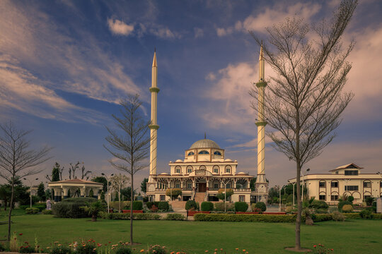 View of Jatoi Mosque with its prominent minarets and dome, set within a well-maintained garden with trees and manicured lawns under a blue sky with soft clouds in Jatoi, Punjab, Pakistan.
