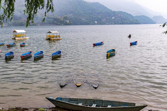View of Phewa Lake with colorful wooden boats and pedal boats floating on the calm water against a backdrop of misty mountains in Pokhara, Gandaki Province, Nepal.