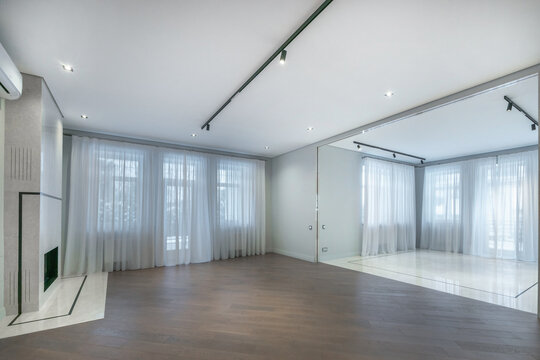 The scene shows a spacious room with large windows and white curtains. A mirrored wall reflects the light. The wooden floor adds warmth to the space. This room can be used for different activities