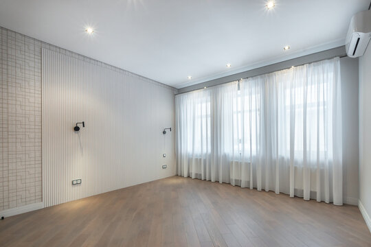 A room with no furniture and wooden floor. There are large windows covered with light curtains. The walls are simple with some textures. It is daytime
