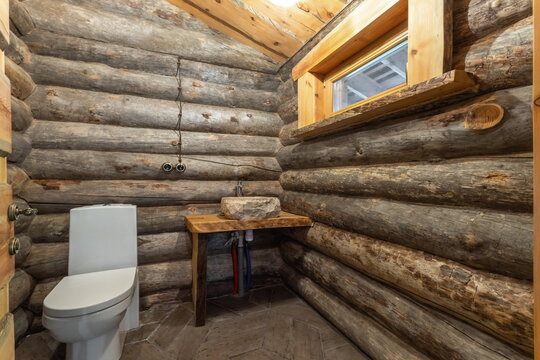 A bathroom inside a log cabin shows a white toilet next to a wooden sink and counter. Natural logs form the walls, and a small window allows light in