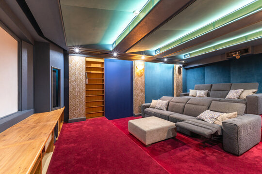 A home theater features gray seating arranged for comfort and viewing. The space includes a large screen, decorative walls, and soft lighting. Shelves are visible in the background