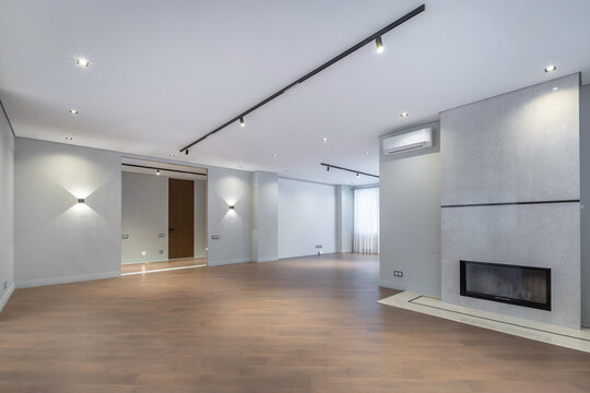 A bare living room shows wooden floors, a fireplace, and walls with lights. Windows let in natural light during the day, creating a bright space for future use