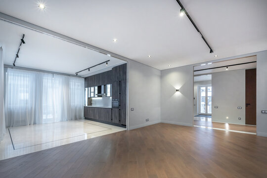 A spacious living area connected to a kitchen in a bright apartment. Large windows allow sunlight to enter. The floor is light wood