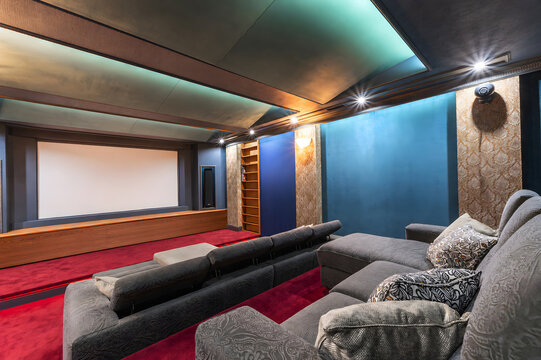 A home theater room features plush seating arranged in front of a large screen. The red carpet and soft lighting create a cozy place for watching films. Shelves hold media items.