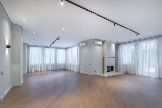 The scene shows an empty living room located in a modern house. Large windows allow light to enter. The wooden floor reflects the bright lights above