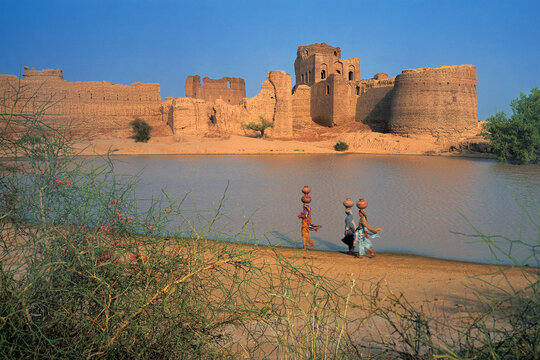 View of Mauj Garh Fort with local women carrying water pots on their heads walking along the shore of a pond in the desert landscape of Mauj Garh, Punjab, Pakistan.