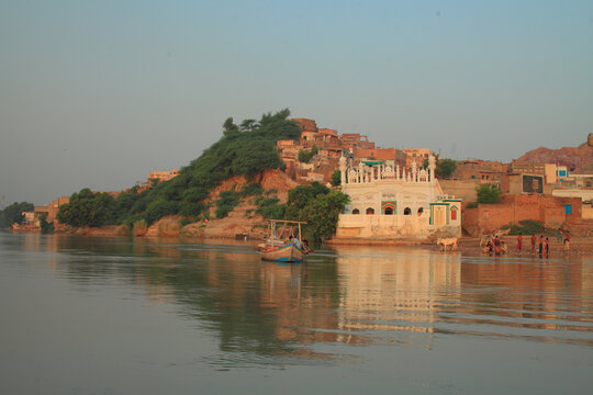 View of the historic town of Kalabagh on the banks of the Indus River, featuring a white mosque, a small wooden boat, and local people along the shore in Kalabagh, Punjab, Pakistan.