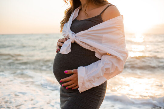 Pregnant woman wearing maternity clothes posing over sea and sunset at beach outdoors. Motherhood. Summer season.