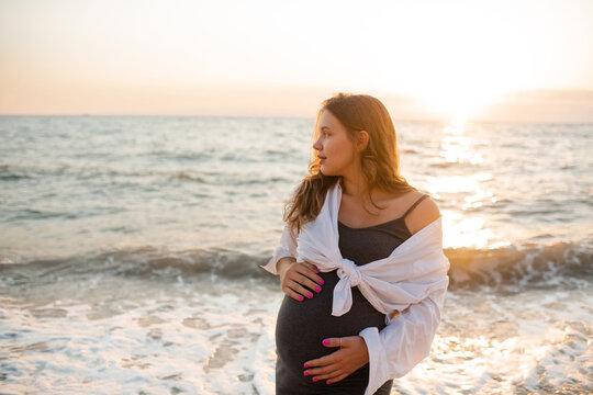 Pregnant woman wearing maternity clothes posing over sea and sunset at beach outdoors. Motherhood. Summer season.