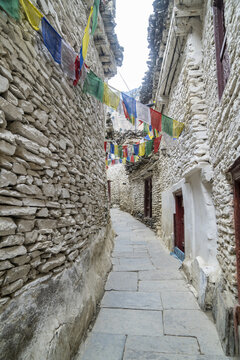 View of a narrow stone-walled alley in Marpha village, adorned with colorful prayer flags, featuring traditional stone architecture and a paved path in Jomsom, Gandaki Province, Nepal.
