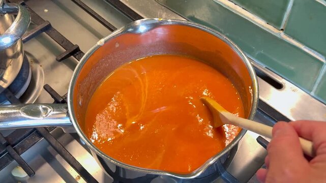 Saucepan of homemade roasted tomato and red pepper soup being stirred while being heated on a hob in a home kitchen, 
