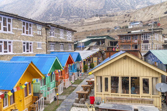 View of Alpine Home guesthouse featuring colorful small wooden cabins with metal roofs, prayer flags, and stone buildings in Tanki Manang, Gandaki Province, Nepal.