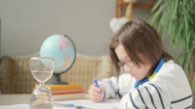 Teenager sitting at desk writing homework in notebook, hourglass time management, education, knowledge and writing, learning process, exam prep, child training, concentration and discipline sand timer
