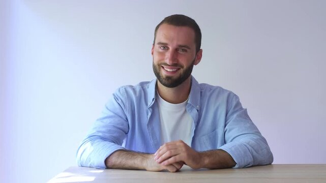 Charming young man with a beard sitting at a table and smiling while looking at the camera, exuding confidence and positivity against a clean, isolated white background in a studio