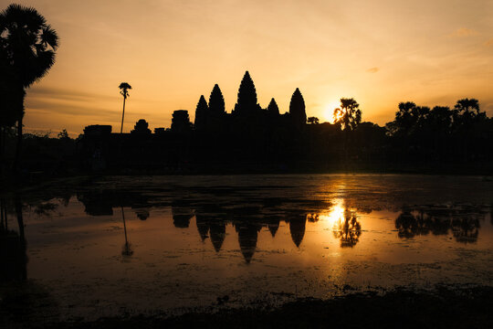 View of the silhouette of Angkor Wat temple at sunrise with its reflection in the water and palm trees in Krong Siem Reap, Siem Reap Province, Cambodia.
