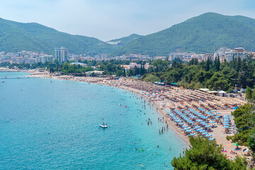 Fototapeta premium Crowded Slovenska Beach stretches along the turquoise bay. Green mountains rise behind the resort city. Budva, Montenegro - June 20, 2021