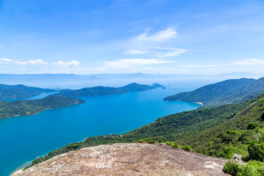 View of Mamangua Bay with lush green hills, deep blue water, and a rocky foreground in Paraty, Rio de Janeiro, Brazil.