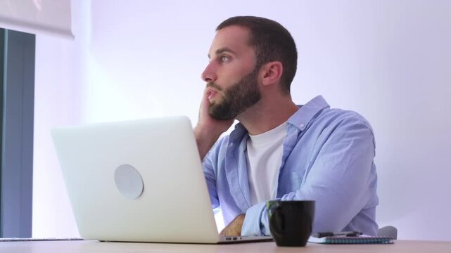 Thoughtful young freelancer working on a difficult project on his laptop computer, feeling stressed and searching for inspiration and new ideas while looking out the window