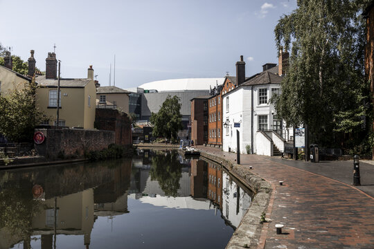 View of the historic canal network with traditional brick buildings, reflections on the calm water, and a modern stadium structure in the background Birmingham, England, United Kingdom.