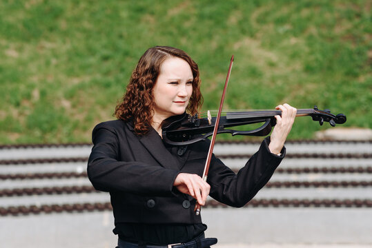 violinist outdoors, young beautiful redhead caucasian woman in jacket playing black violin in city park with green grass and trees in sunny summer day, classic music concept