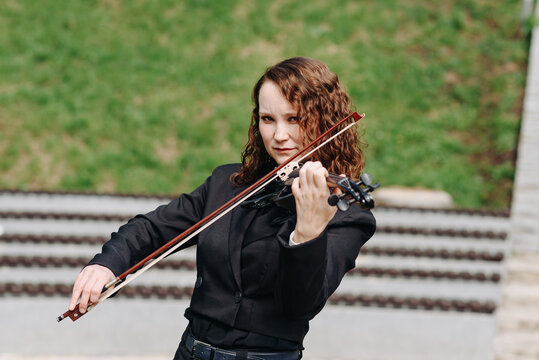 violinist outdoors, young beautiful redhead caucasian woman in jacket playing black violin in city park with green grass and trees in sunny summer day, classic music concept