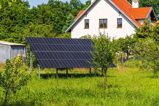 View of solar panels installed in a lush green garden with a house in the background in Kadyny, Warmian-Masurian Voiv, Poland.