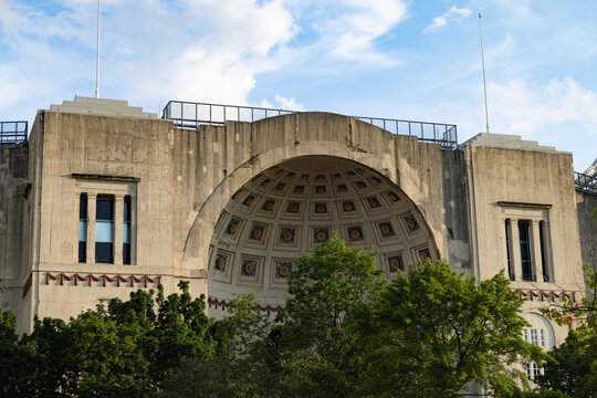 View of the historic rotunda and arched entrance of Ohio Stadium surrounded by lush green trees under a blue sky in Columbus, Ohio, United States.